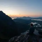 A hiker sitting on a rocky outcrop on Nonstinden in Ballstad, Lofoten, enjoying a peaceful sunset view over the fjords, lakes, and surrounding mountains.