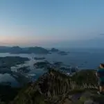 A hiker stands on the summit of Nonstinden in Ballstad, Lofoten, overlooking the town and surrounding fjords at sunset.