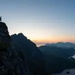 A hiker standing on the edge of a rocky cliff at sunset on Nonstinden in Ballstad, Lofoten, with panoramic views of fjords, lakes, and distant mountain peaks.
