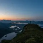 A hiker standing on the summit of Nonstinden in Ballstad, Lofoten, enjoying a breathtaking sunset view over the fjords, lakes, and surrounding mountain peaks.