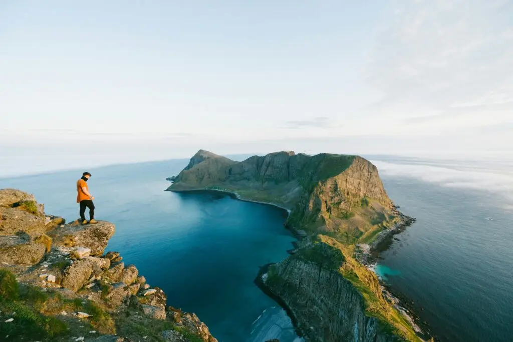 A hiker standing on the summit of Håen (Håheia) at 438m on Værøy, Lofoten, overlooking the dramatic coastline, turquoise waters, and steep cliffs.