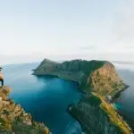 A hiker standing on the summit of Håen (Håheia) at 438m on Værøy, Lofoten, overlooking the dramatic coastline, turquoise waters, and steep cliffs.