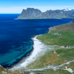 Aerial view of Uttakleiv Beach with turquoise waters, green meadows, and dramatic mountain peaks, seen from the Veggen hike in Lofoten, Norway.