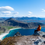 Person sitting on a mountain peak overlooking Haukland and Vik Beaches with turquoise waters and dramatic mountain views in Lofoten, Norway.