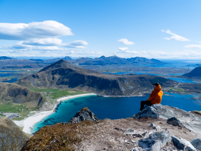 Person sitting on a mountain peak overlooking Haukland and Vik Beaches with turquoise waters and dramatic mountain views in Lofoten, Norway.