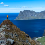 Hiker in orange jacket standing on a mountain ridge above Uttakleiv Beach with dramatic ocean and mountain views in Lofoten, Norway.