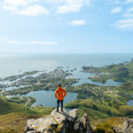 Hiker standing on Tuva summit in Lofoten, overlooking Svolvær and the Vestfjord on a sunny day.