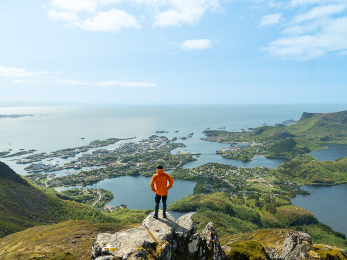 Hiker standing on Tuva summit in Lofoten, overlooking Svolvær and the Vestfjord on a sunny day.