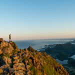 Hiker standing on rocky summit of Blåtinden in Lofoten at sunset, overlooking sea and mountain landscape