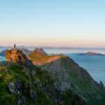 Person standing on rocky outcrop on Blåtinden in Lofoten during golden hour, with wide view of mountains and sea