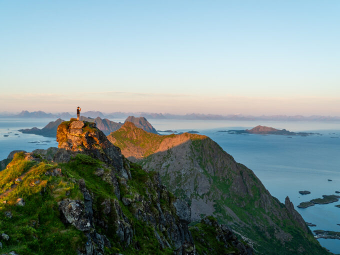 Person standing on rocky outcrop on Blåtinden in Lofoten during golden hour, with wide view of mountains and sea