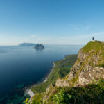 View from Hornet on Værøy with hiker on cliff edge, Mosken and Lofoten mainland in the background