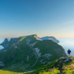 Hiker on Hornet summit, Værøy, with winding military road and steep cliffs in the background