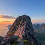 A hiker stands on the rocky summit of Moltinden in Lofoten as the sky glows at sunset.